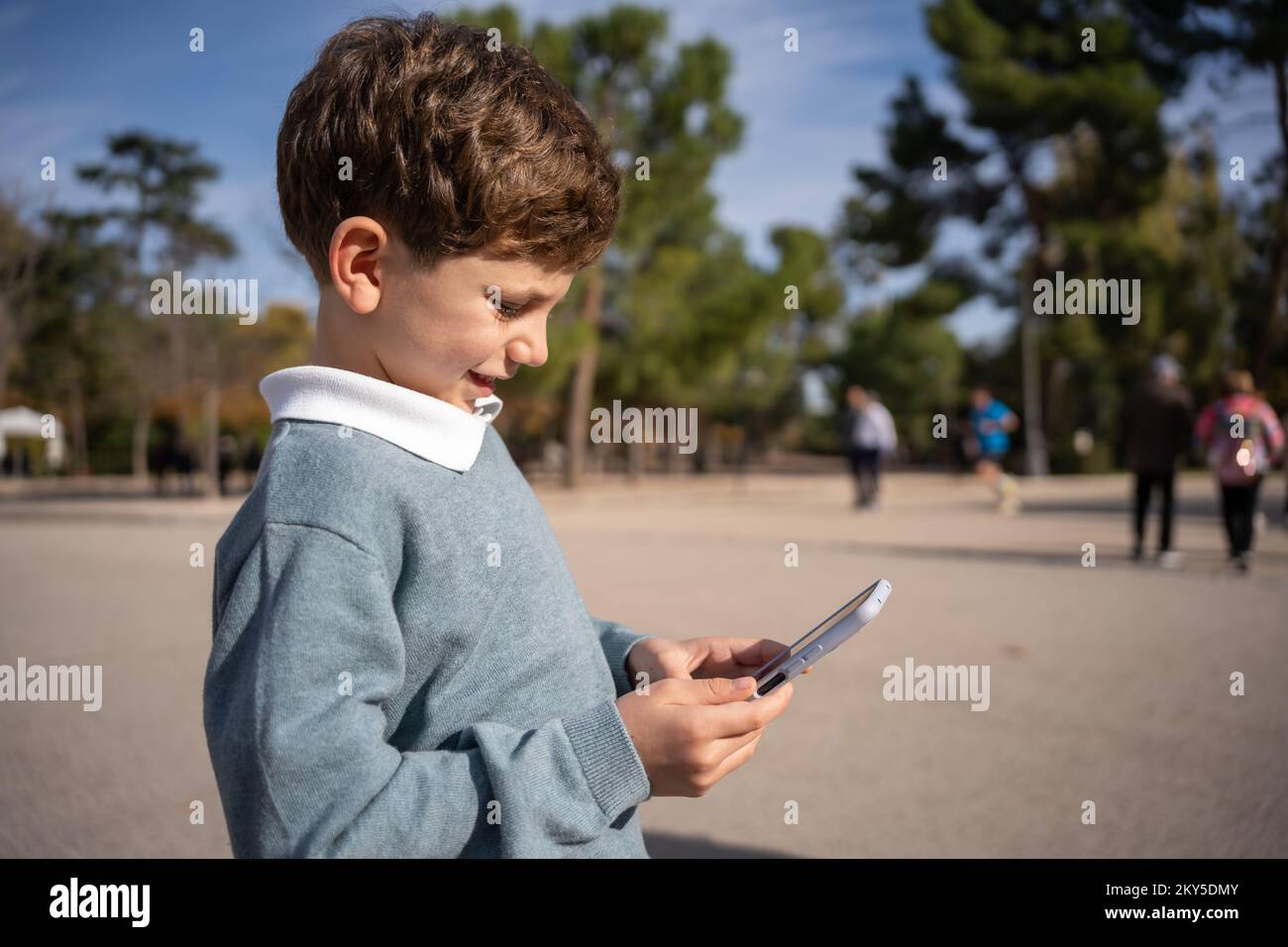 A dark-haired boy holds a smart phone as he plays quietly while standing on a street with green trees in the background Stock Photo