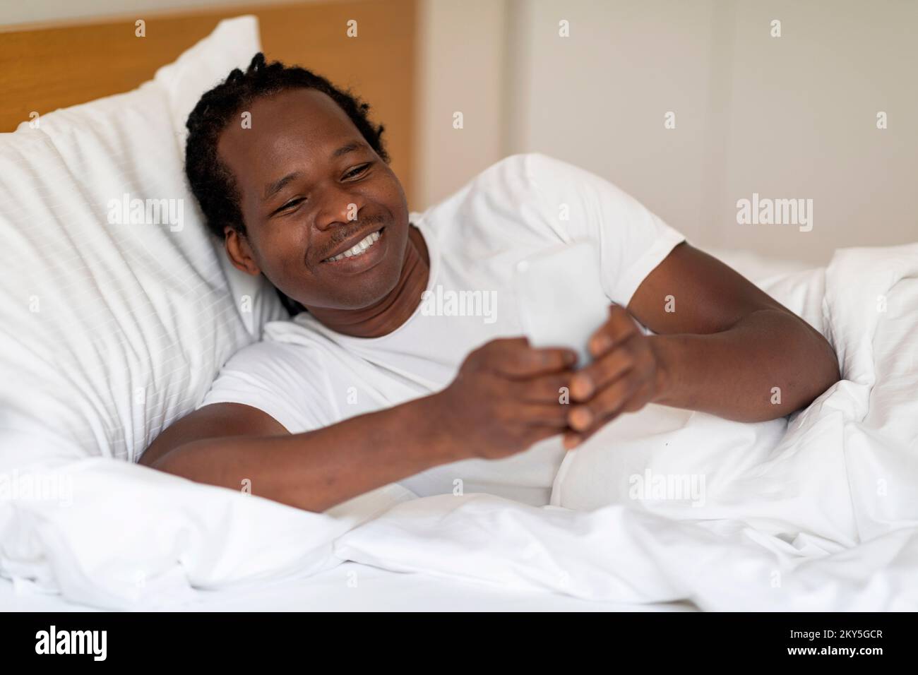 Smiling Black Man Playing Online Games On Smartphone While Lying In Bed Stock Photo