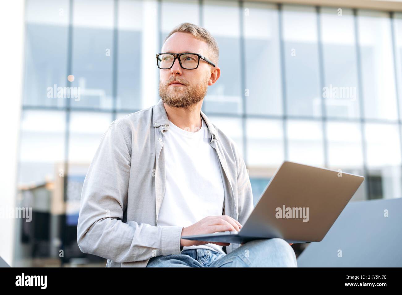 Serious focused caucasian bearded man with glasses, in casual clothes, sits outdoors near the office with a laptop, works on a project, looks away thoughtfully, plans a strategy, thinking over idea Stock Photo