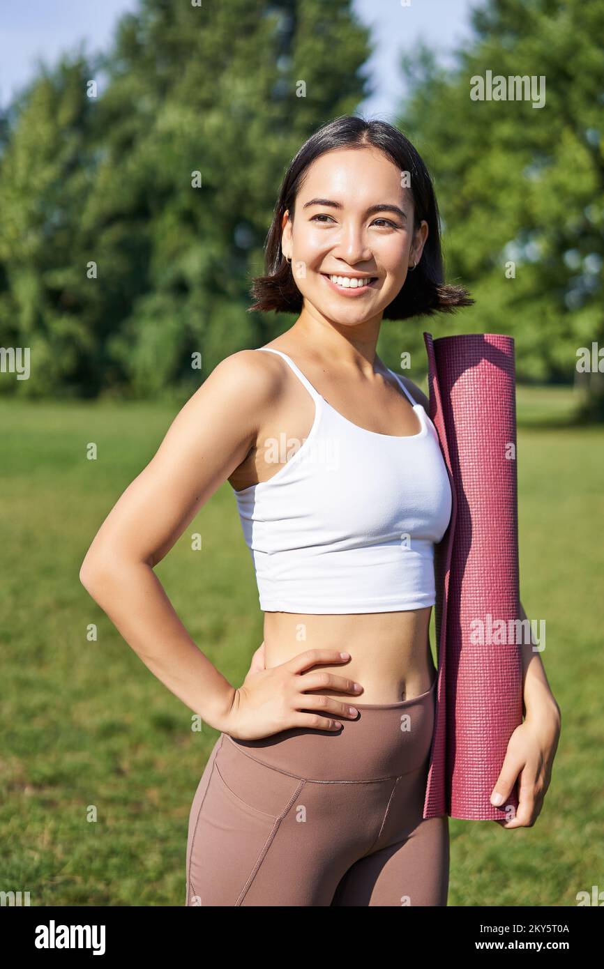Smiling fitness girl with rubber mat, stands in park wearing uniform