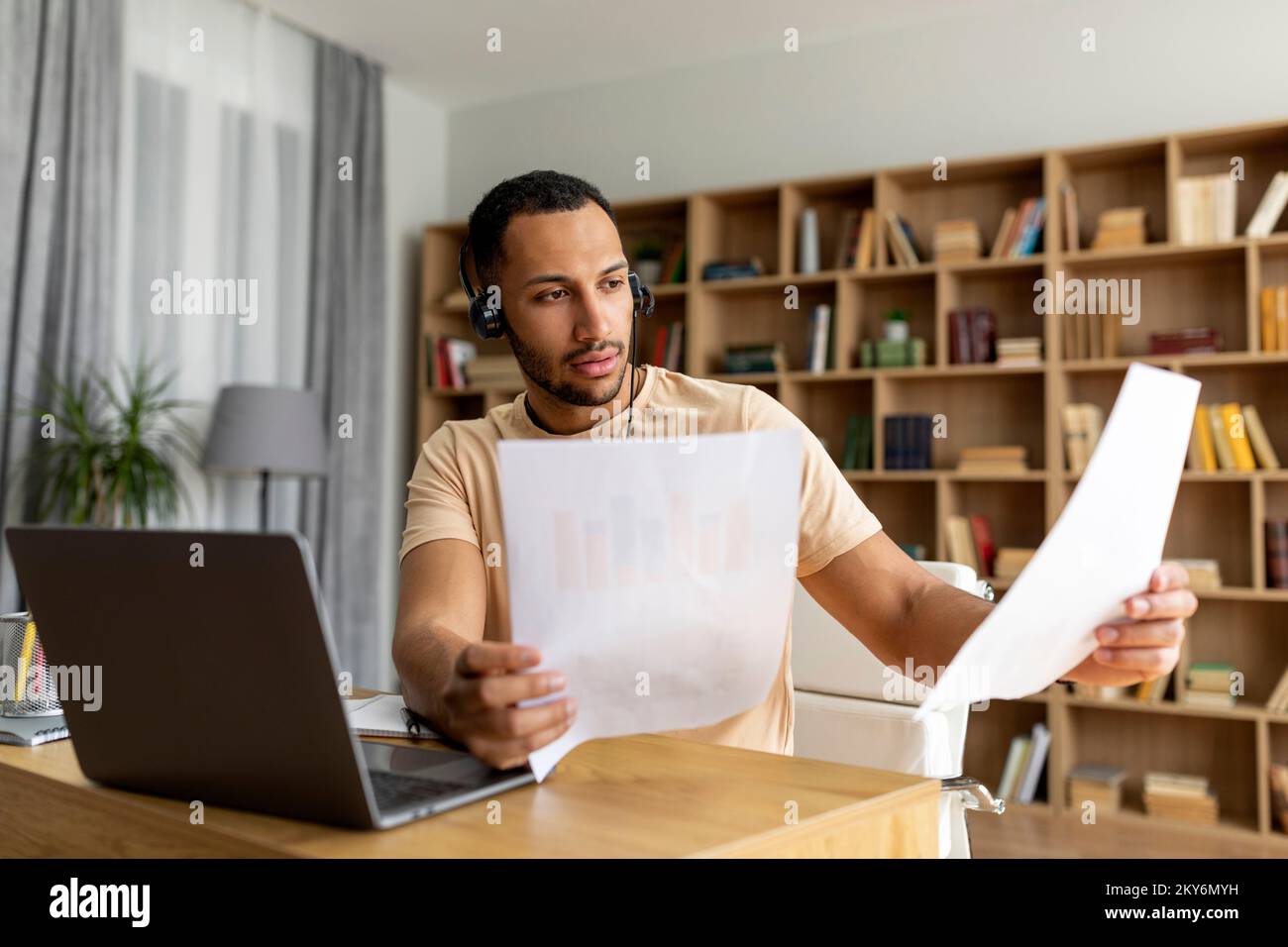 Focused arab man in wireless headphones having video call on laptop, working with documents at home in living room Stock Photo