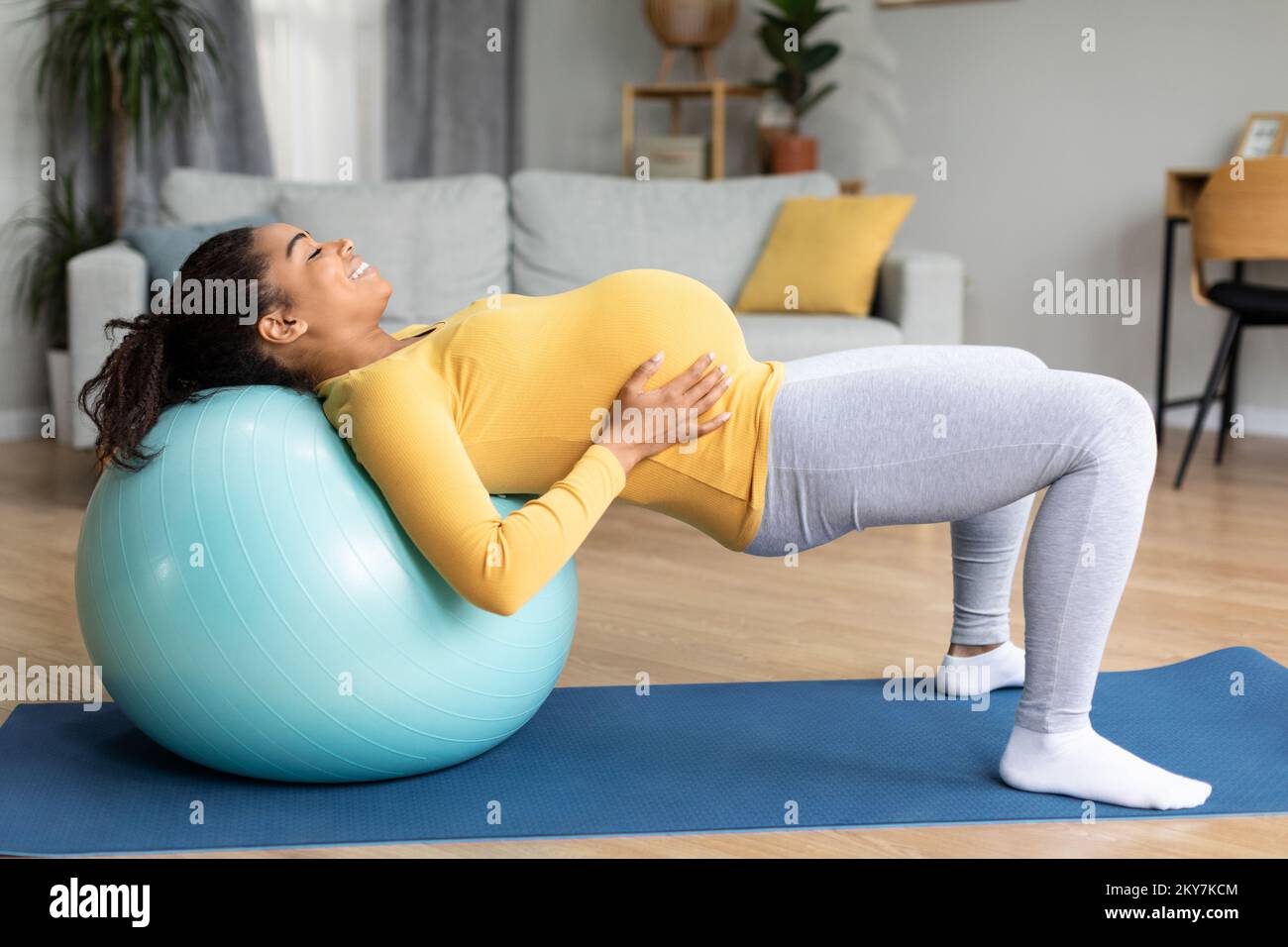 Happy young black pregnant lady with big belly doing exercises on fitness ball, feels baby