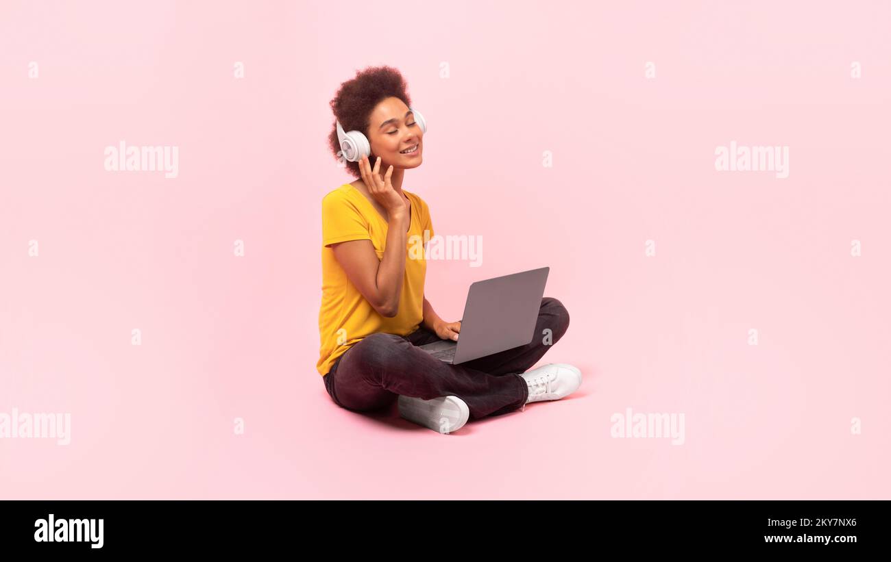 Happy millennial african american curly woman student in wireless headphones sit on floor with laptop enjoy music Stock Photo