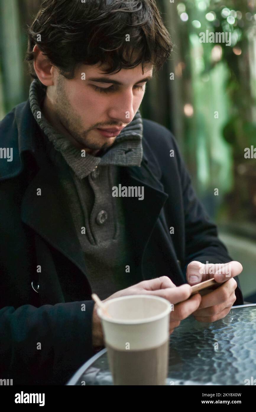 Young handsome man in coat with coffee texting on cellphone Stock Photo