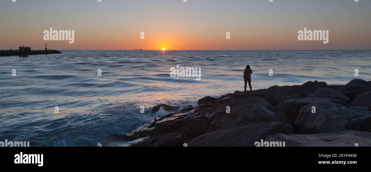 Sun setting on sea horizon with waves lapping against foreground rocks and a silhouetted human figure watching the scene with a distant merchant ship Stock Photo