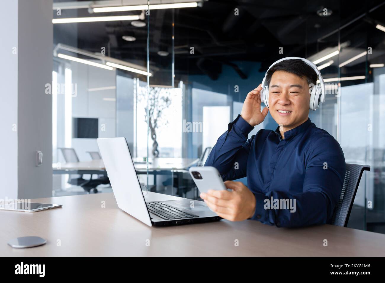 Businessman listening to music and podcast at workplace, asian man smiling and happy using laptop at work, man with headphones and phone listening to online radio inside office. Stock Photo