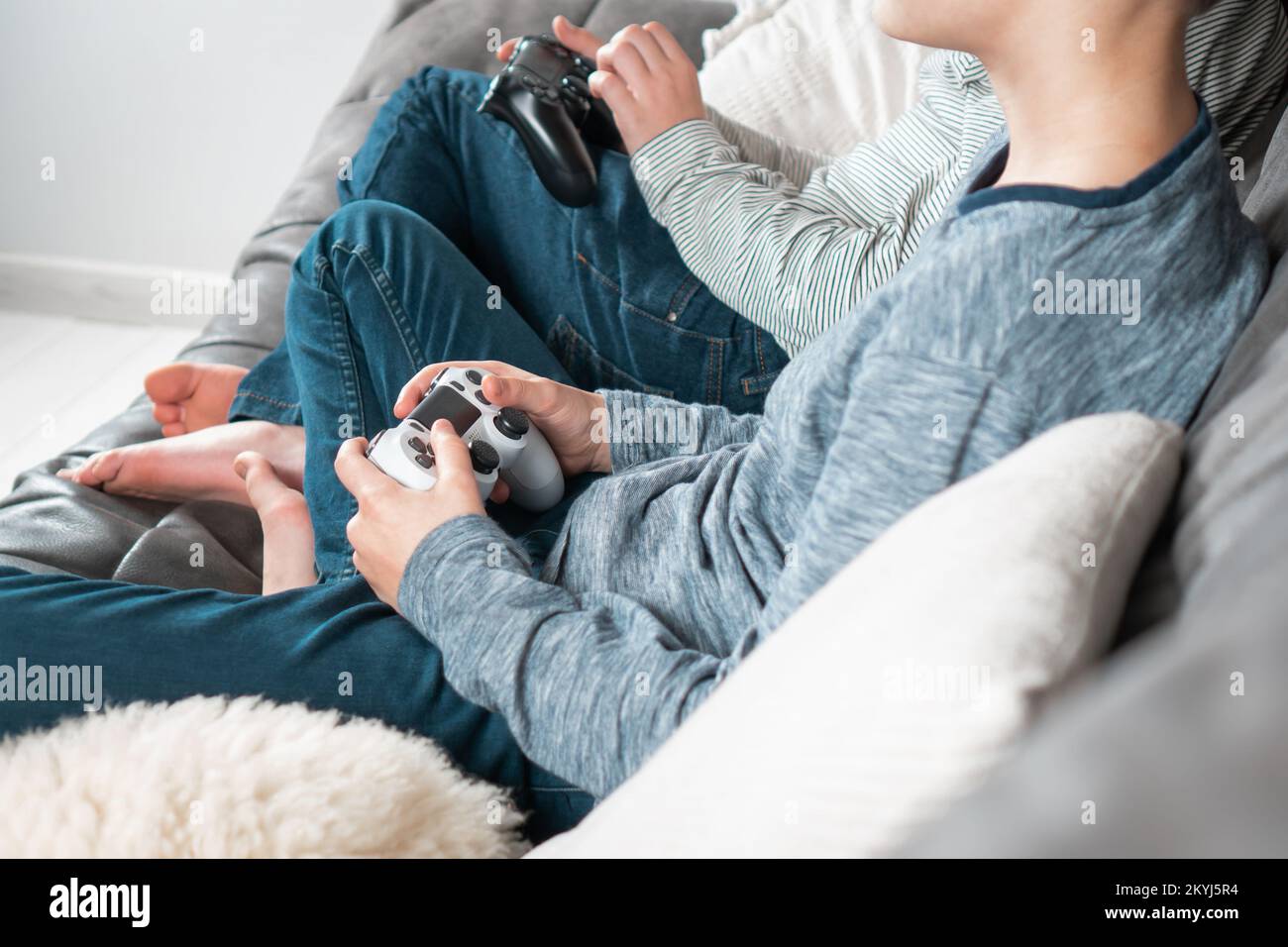 Hands of guys sitting on sofa with game joysticks, side view. Children play videogames. Boys in denim and casual longsleeves relaxing at home Stock Photo