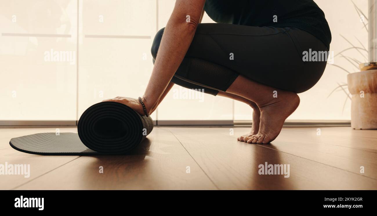 Unrecognisable senior woman rolling up an exercise mat after a yoga session. Mature woman following a healthy wellness routine at home. Elderly woman Stock Photo