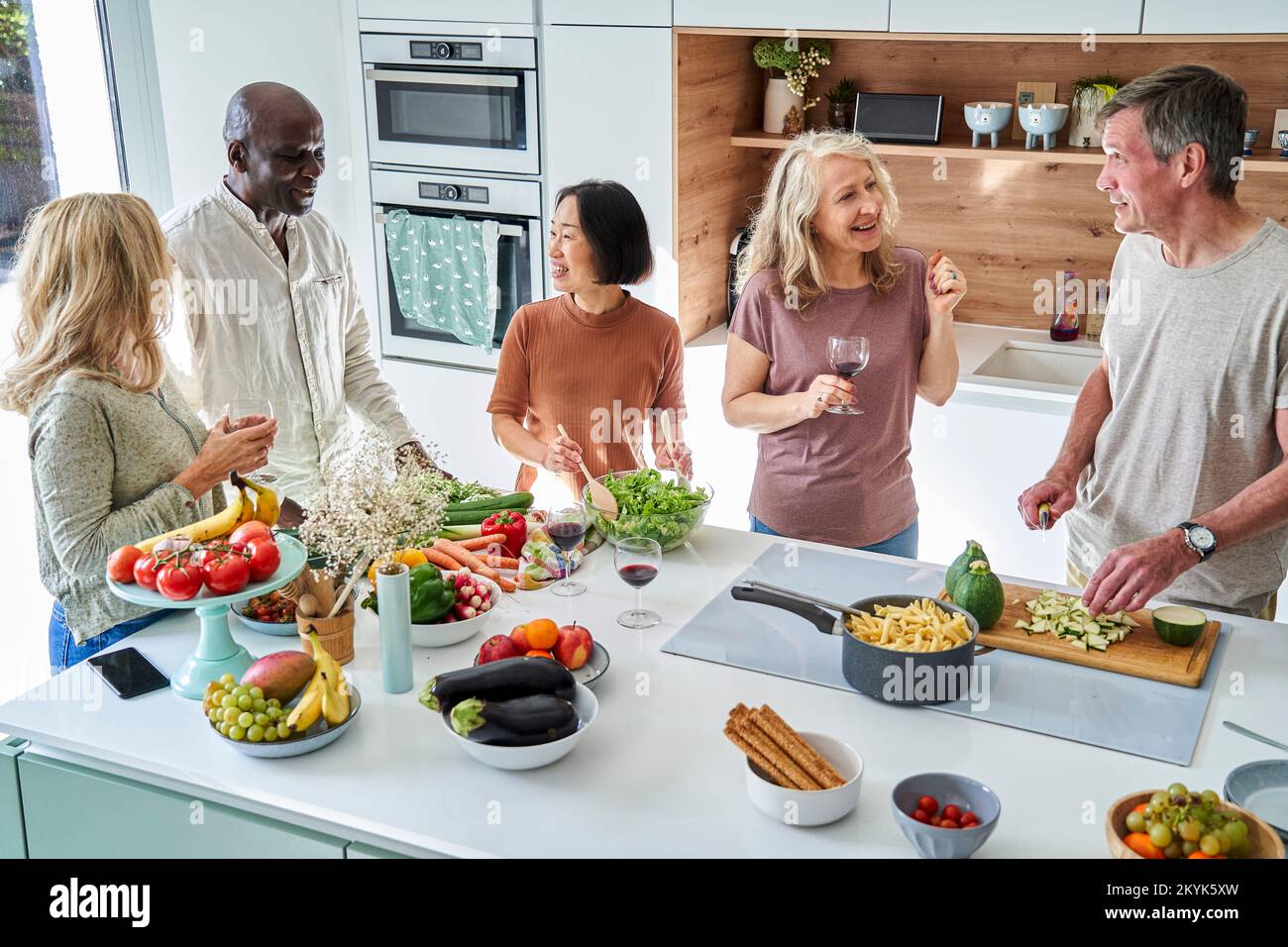 Diverse group of senior friends drinking wine while gathered at kitchen Stock Photo