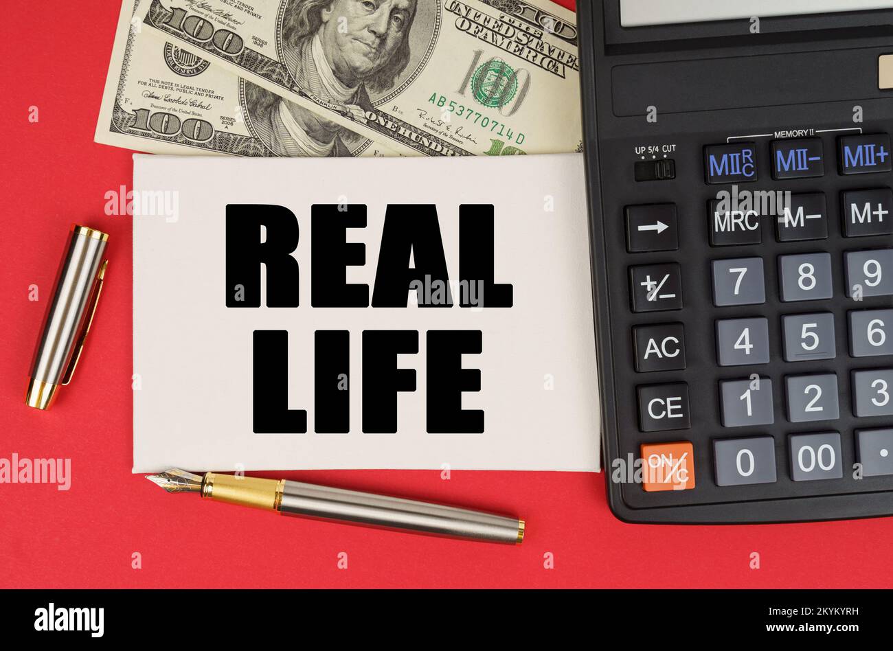Business and finance concept. On a red background, among the money, a calculator and a pen lies a sign with the text - REAL LIFE Stock Photo