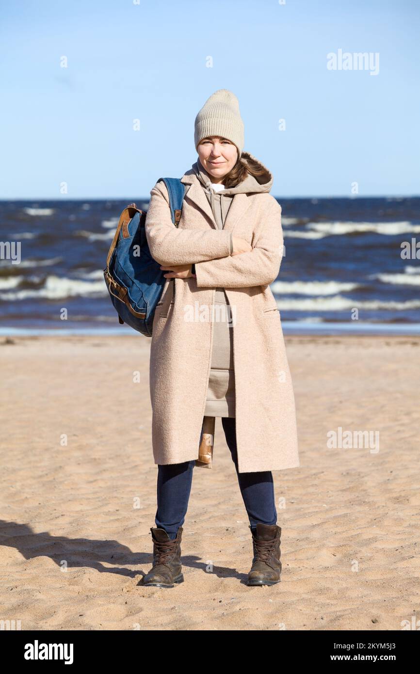 A woman in a coat and with a backpack on her shoulder stands on a sandy