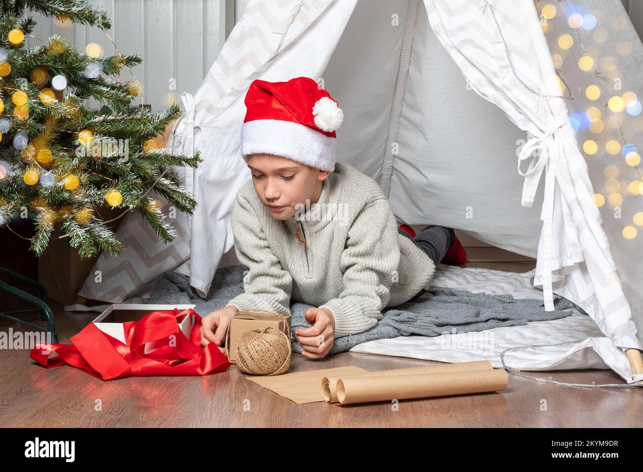 A child in a red Santa hat wraps Christmas gifts surprises in kraft paper with a rope, red ribbons. A teen boy lying in a children's tent is packing g Stock Photo