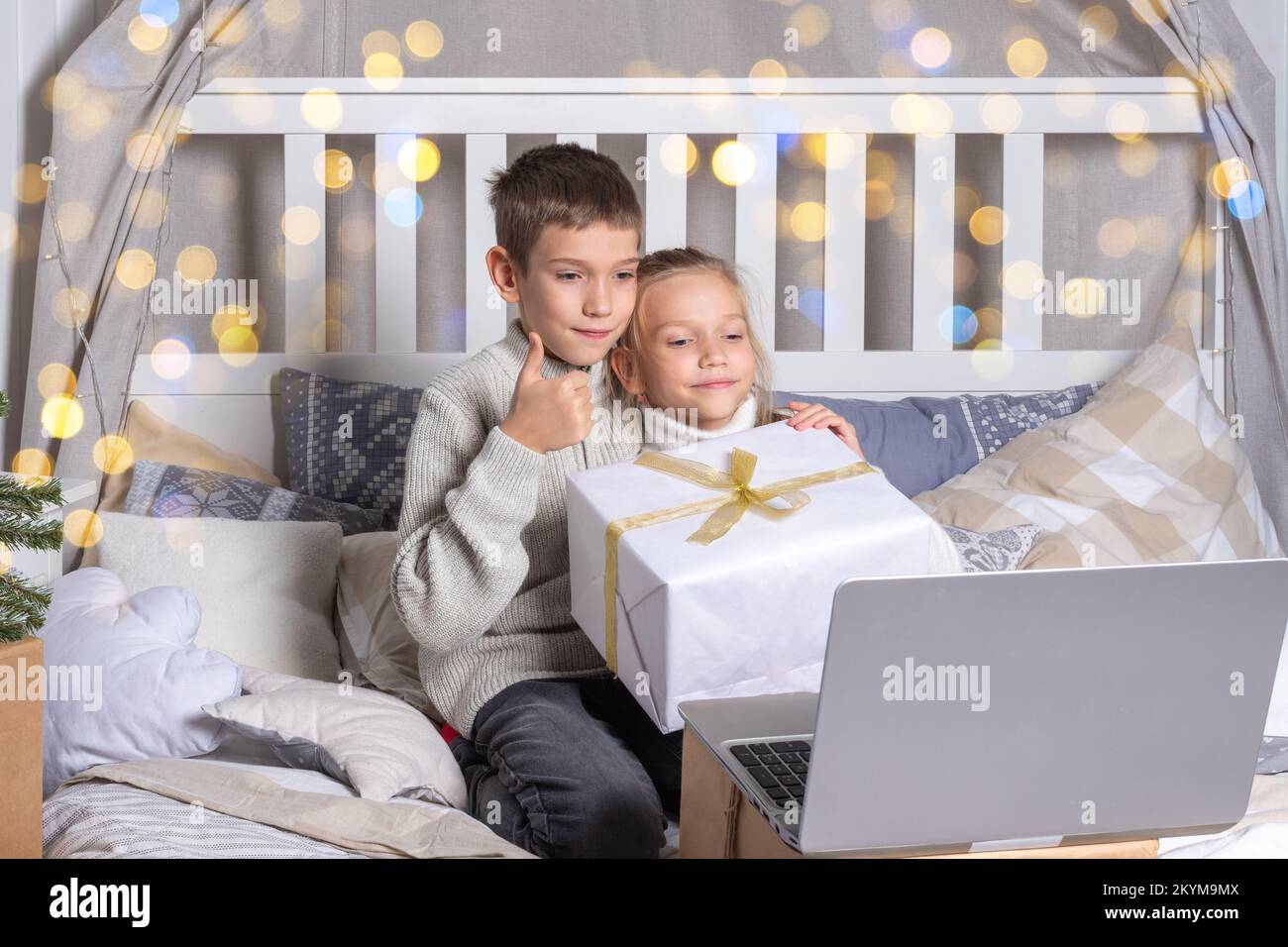 Preschool children holding gift box chatting via video link via laptop and waving hand greeting relatives or friends at home. Boy and girl brother and Stock Photo