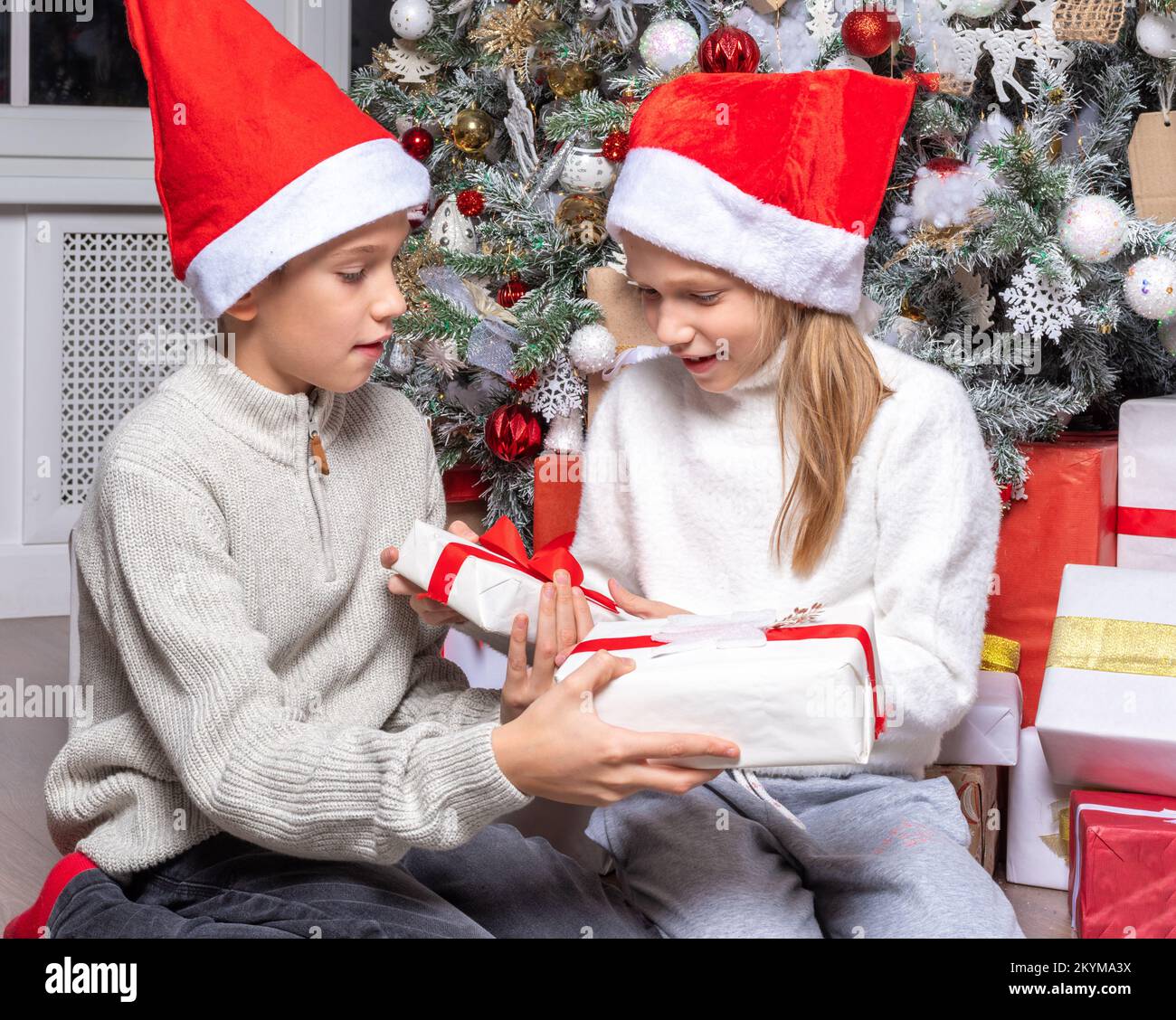 Cute happy excited kids, boy and girl in santa hats exchange surprise gifts next to christmas tree at home. Sister and brother giving Christmas gifts Stock Photo