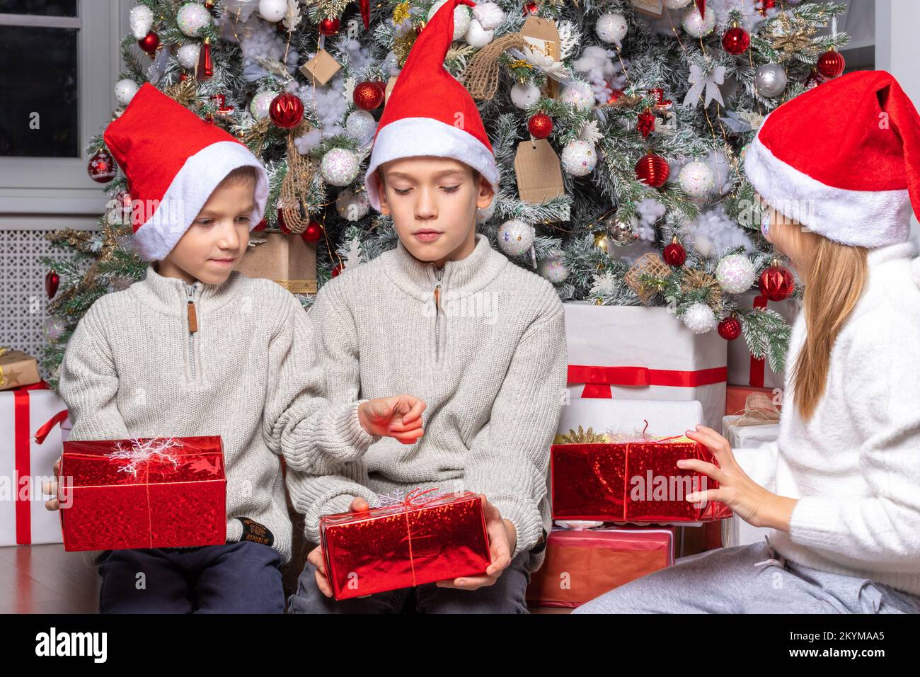 Cute happy excited children, boy and girl in santa hats unwrap christmas present box in beautiful room. Sister and brother give Xmas gifts near decora Stock Photo