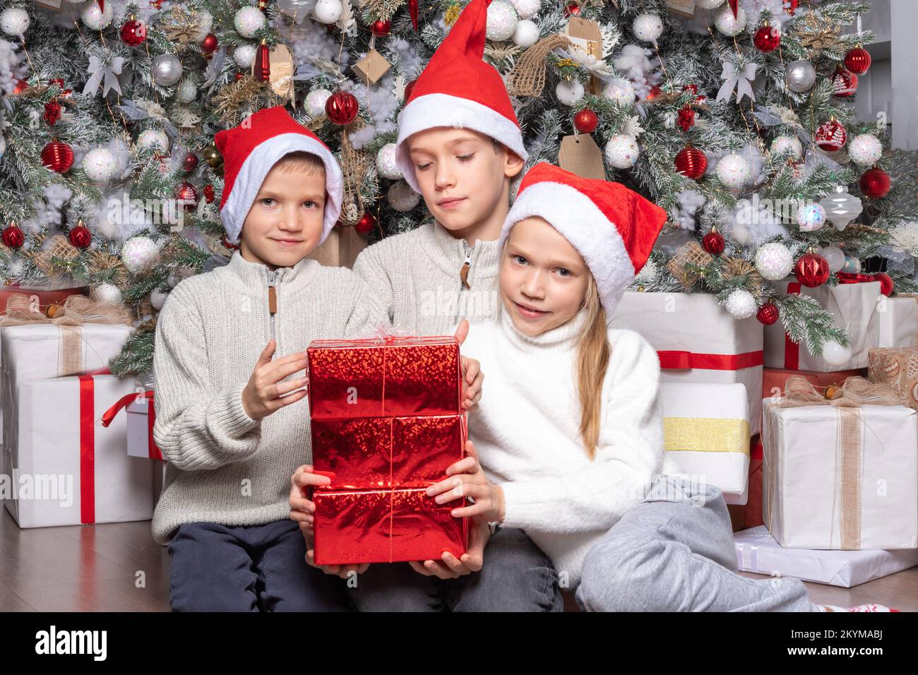 Three cute happy excited kids, boys and a girl in santa hats exchange surprise gifts next to the christmas tree at home. Sister and brothers give Chri Stock Photo