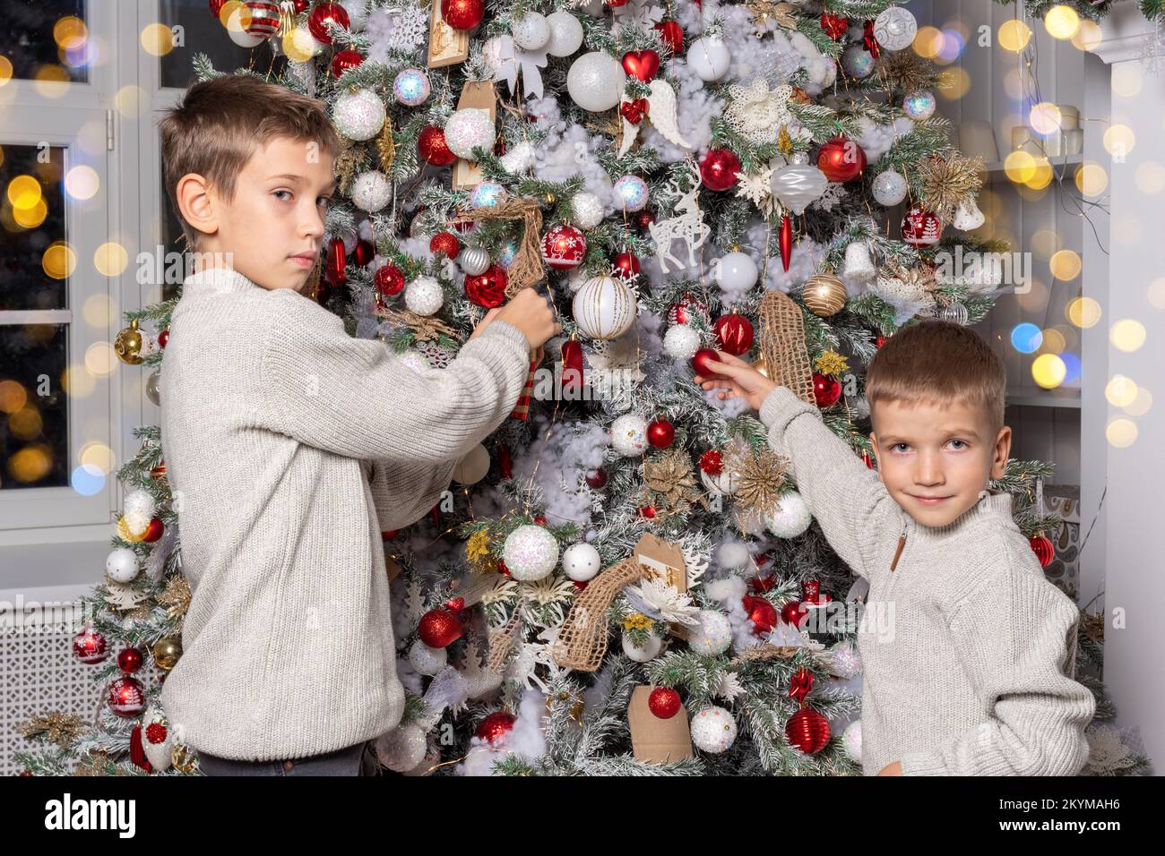 Cute cheerful children, boys decorate a Christmas tree, hang balls, garlands. The brothers decorate the house for the New Year and Christmas holidays. Stock Photo