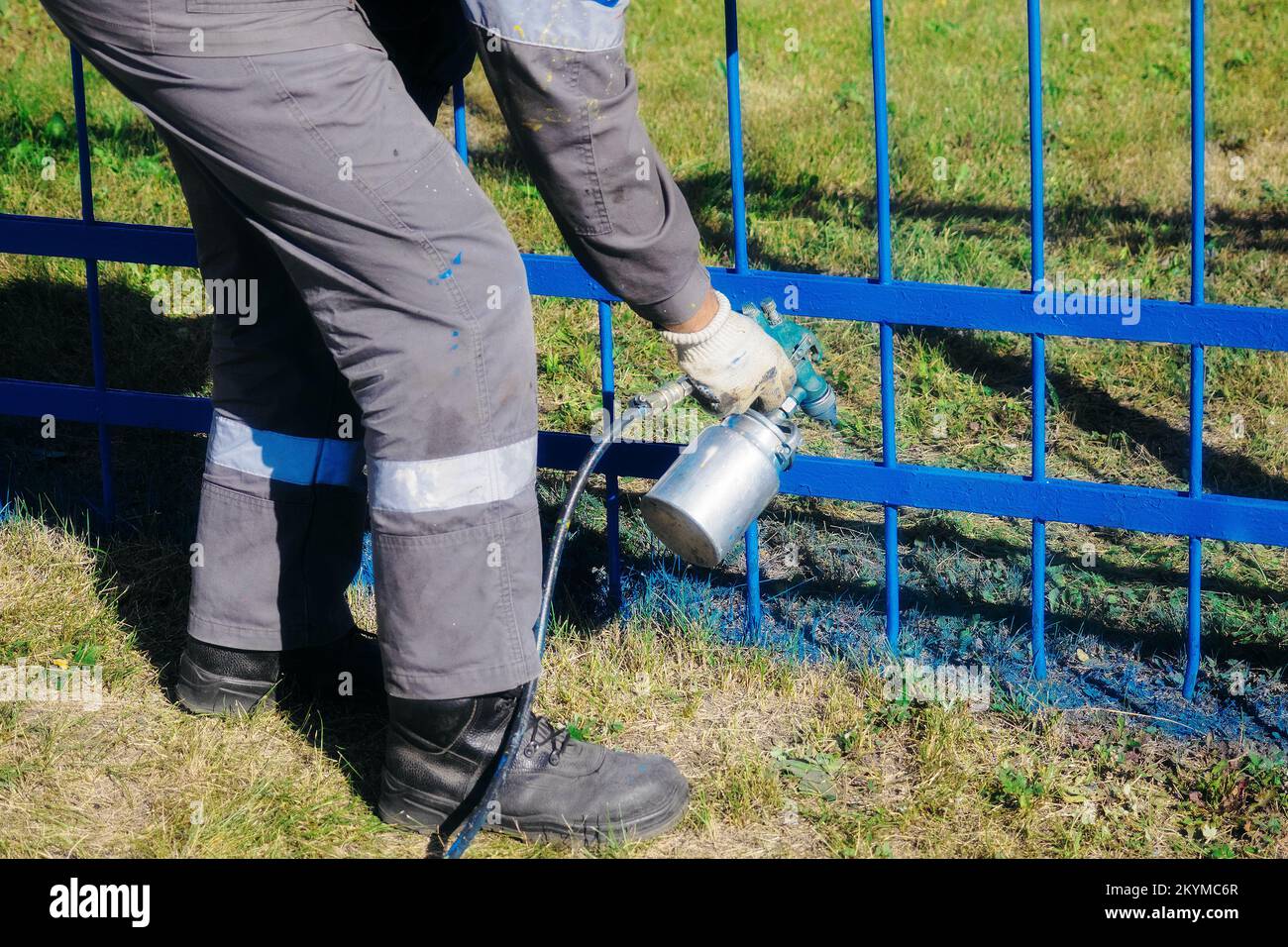 Worker in overalls paints external metal fence from spray gun on summer day. Kind of spray gun