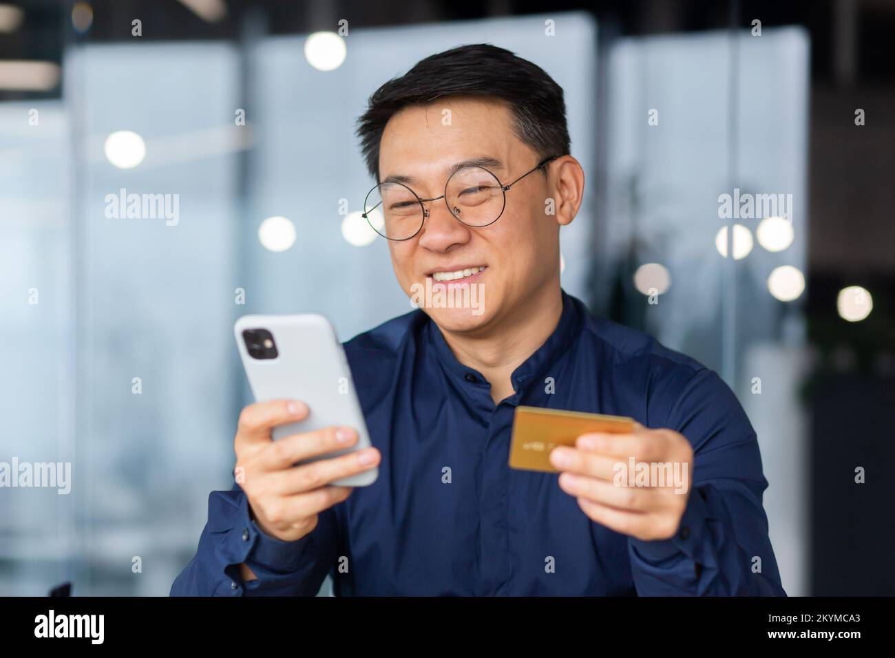 Successful and smiling asian businessman in office making bank transfer and online purchase in online store, man using app on phone and bank credit card, close up inside. Stock Photo