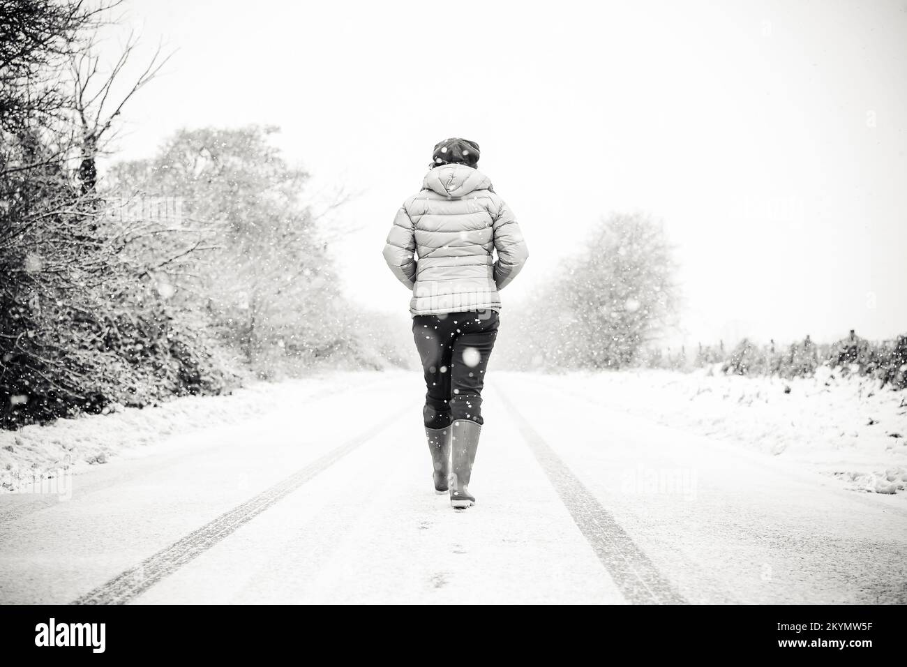 Black & white rear view of a woman wearing winter coat & wellies walking isolated down the middle of a  UK country road in the snow. Stock Photo