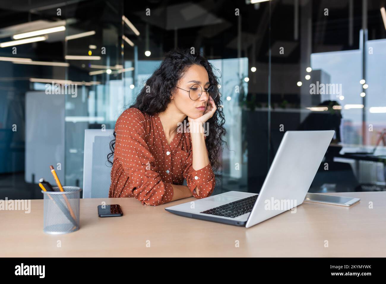 Sad grumpy Latin American woman working inside the office, business woman is bored looking at the laptop screen. Stock Photo