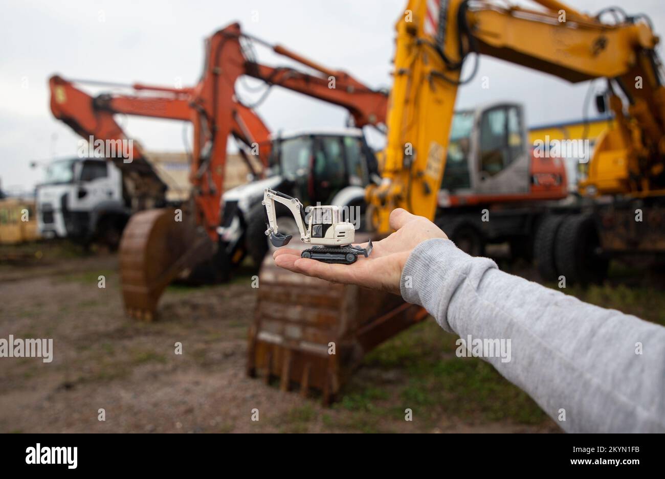 man's hand holds a small toy excavator in his palm. Behind him, out of focus, row of real
