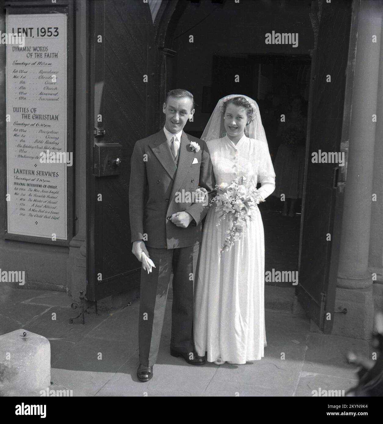 1953, historical, Lent and a newly married couple standing together outside the wooden doors at the entrance to a church, England, UK. Sign on the wall says LENT, 1953 and is enscribed with; 'Dynamic Words of Faith' Duties of the Christian, including Discipline, Giving, Bible Reading, Upholding Christ's ? of Marriage, Worship, Lantern Services, including Personal Interviews of Christ. Stock Photo