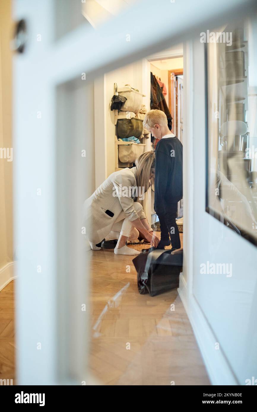 Side view of woman kneeling on floor and assisting son in wearing shoe seen through door Stock Photo