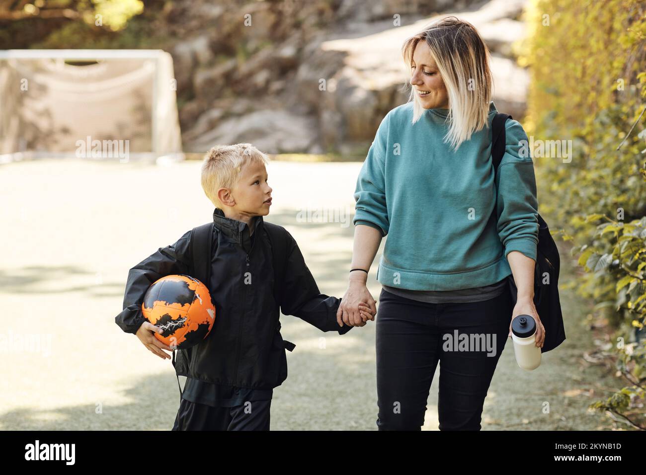 Mother holding hand of son carrying soccer ball while walking in ground Stock Photo