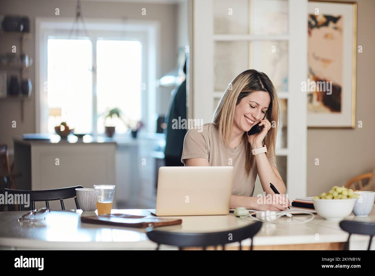 Happy freelancer taking down notes while talking on smart phone at home Stock Photo