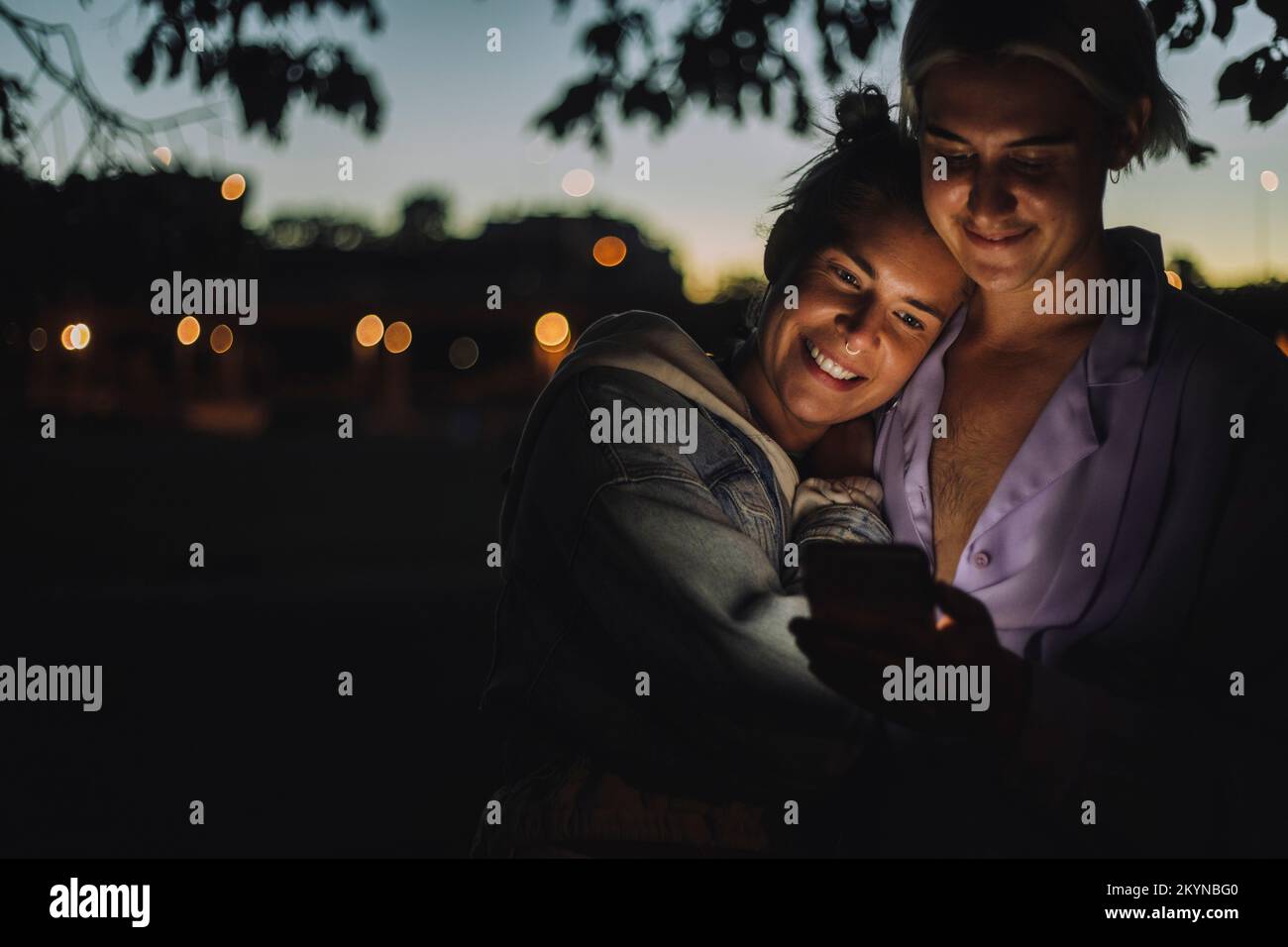 Smiling woman embracing non-binary friend using mobile phone at night Stock Photo