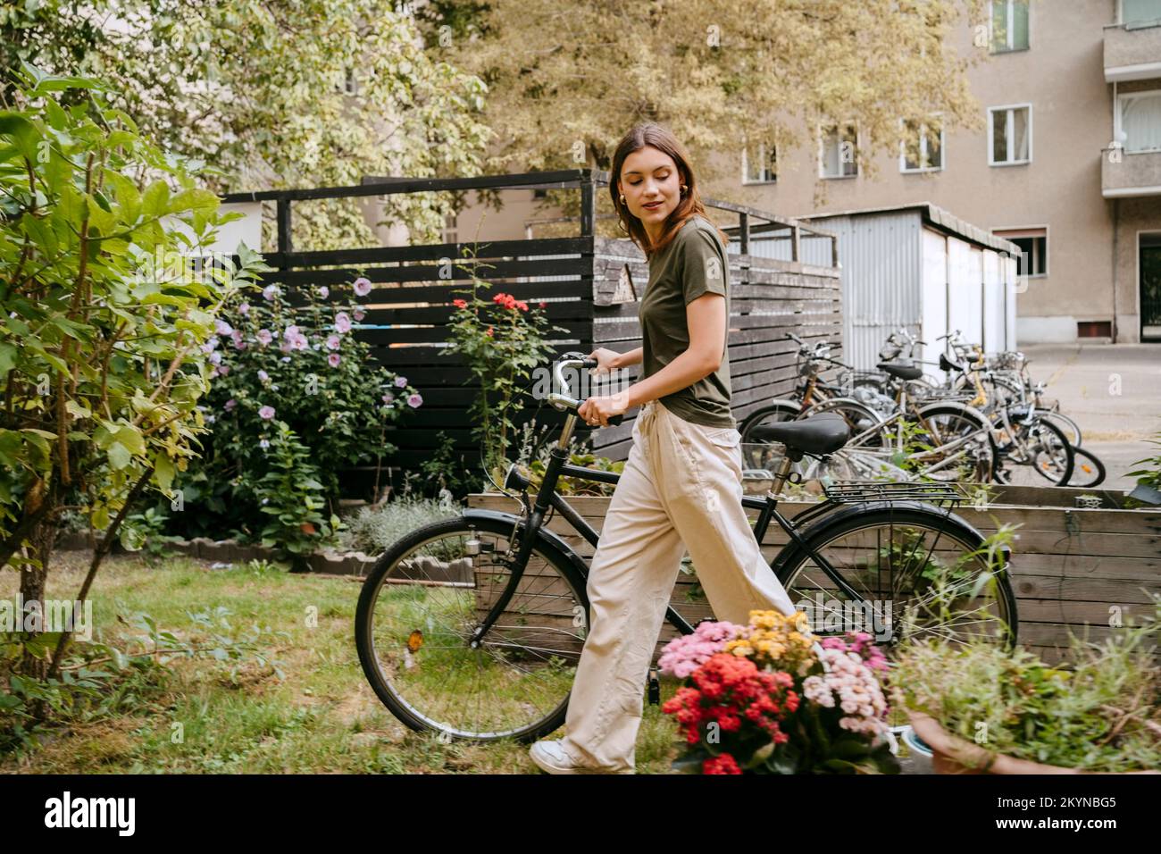 Side view of young woman wheeling with bicycle while looking at flowers in garden Stock Photo