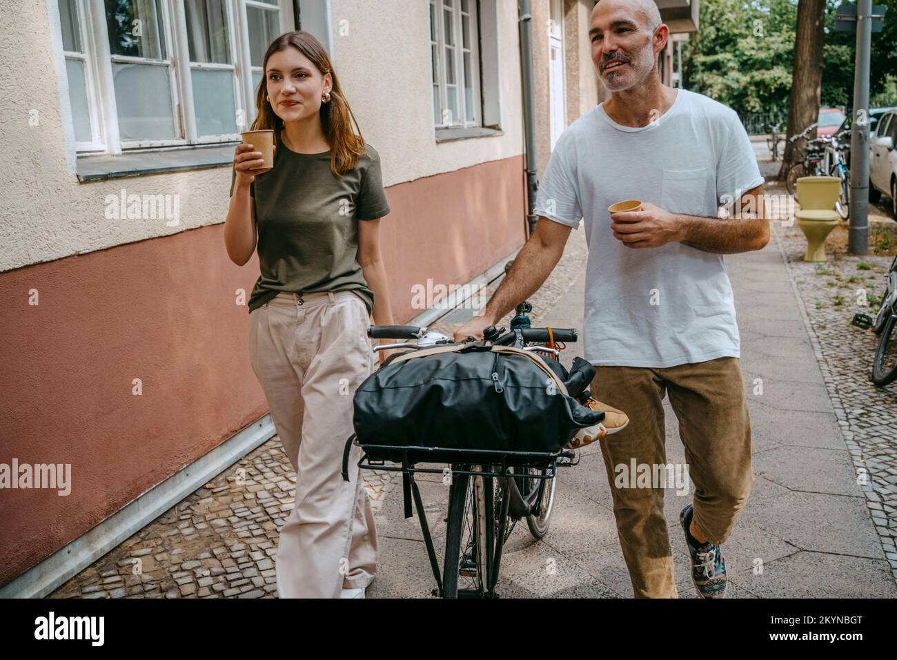 Male and female friends having coffee while wheeling with bicycle on sidewalk Stock Photo