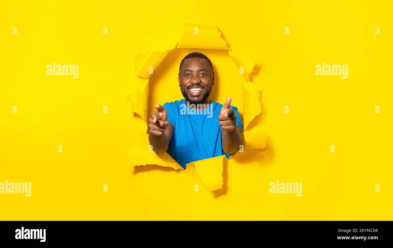 And what's about you. Happy african american man pointing fingers at camera, breaking through hole in yellow paper Stock Photo