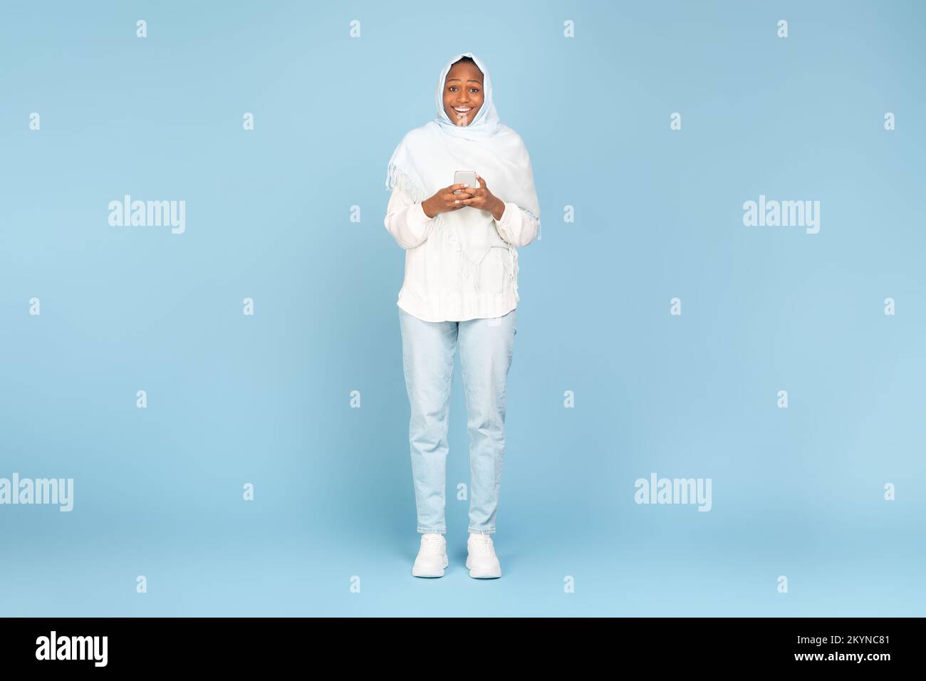 Excited black muslim lady in hijab using smartphone, texting and smiling to camera, standing over blue studio background Stock Photo