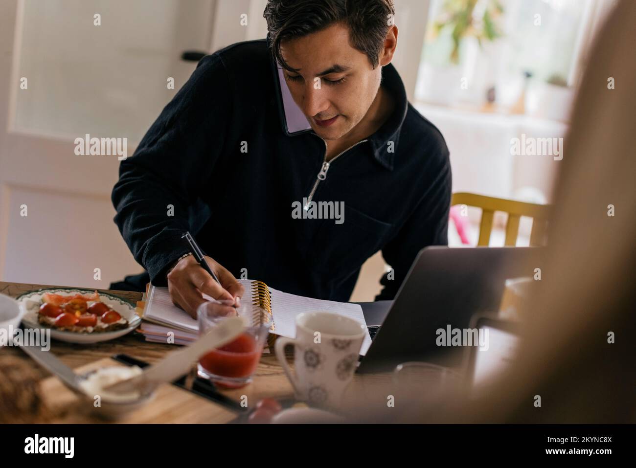 Young male freelancer taking down notes while talking on smart phone at dining table Stock Photo