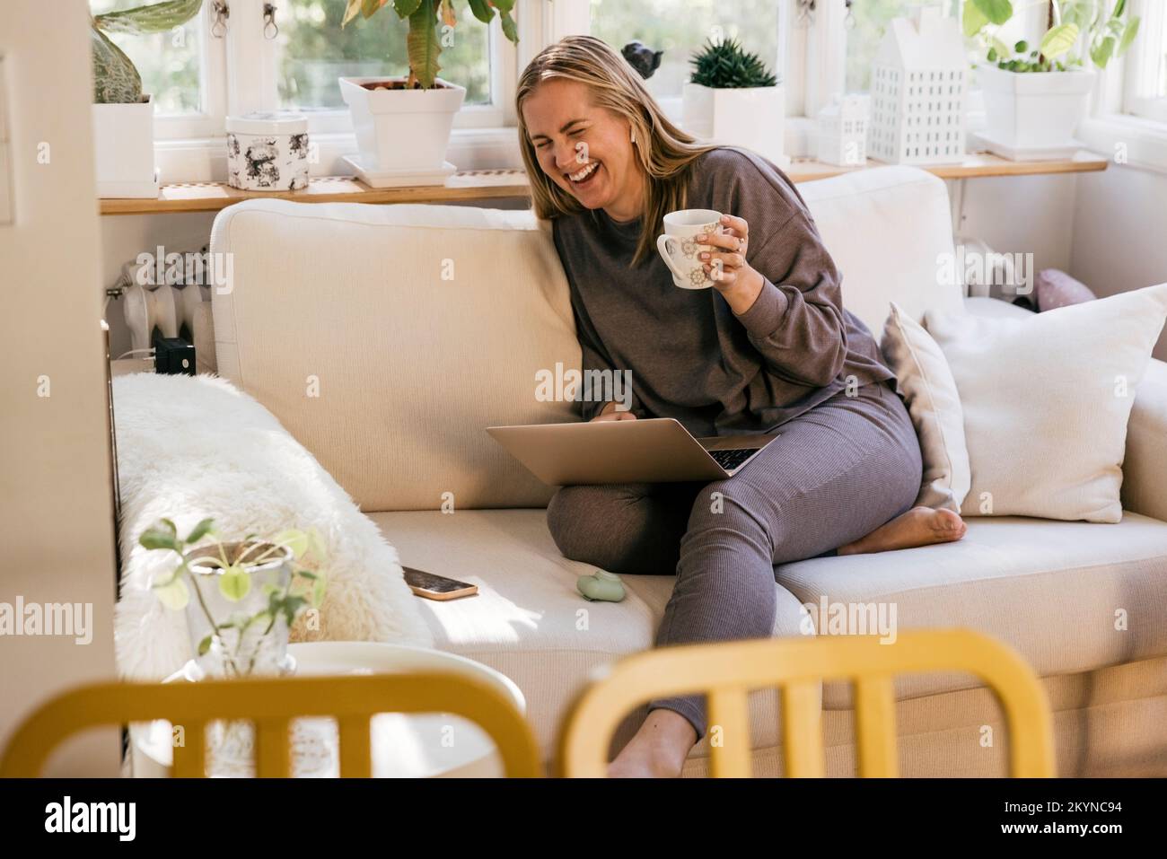 Happy female freelancer with laptop holding mug while sitting on sofa at home Stock Photo