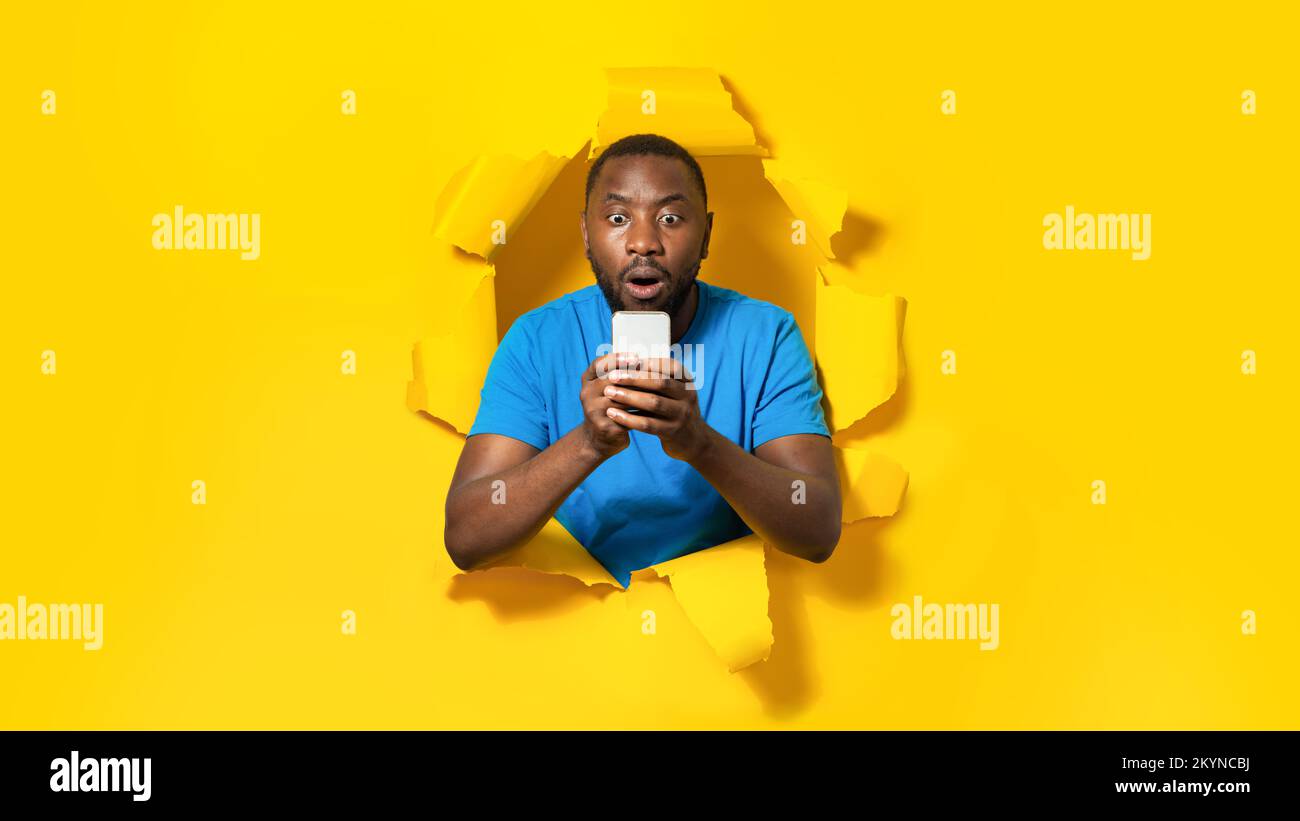 Shocked black man using cellphone, reading shocking message, posing in hole of torn paper on yellow background Stock Photo