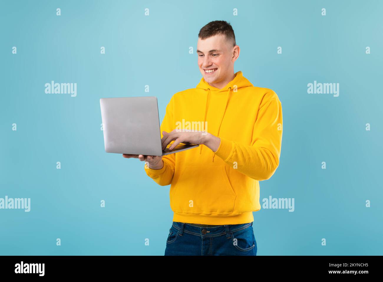 Happy handsome caucasian man enjoying his new modern laptop, typing on keyboard and smiling, blue background, copy space Stock Photo