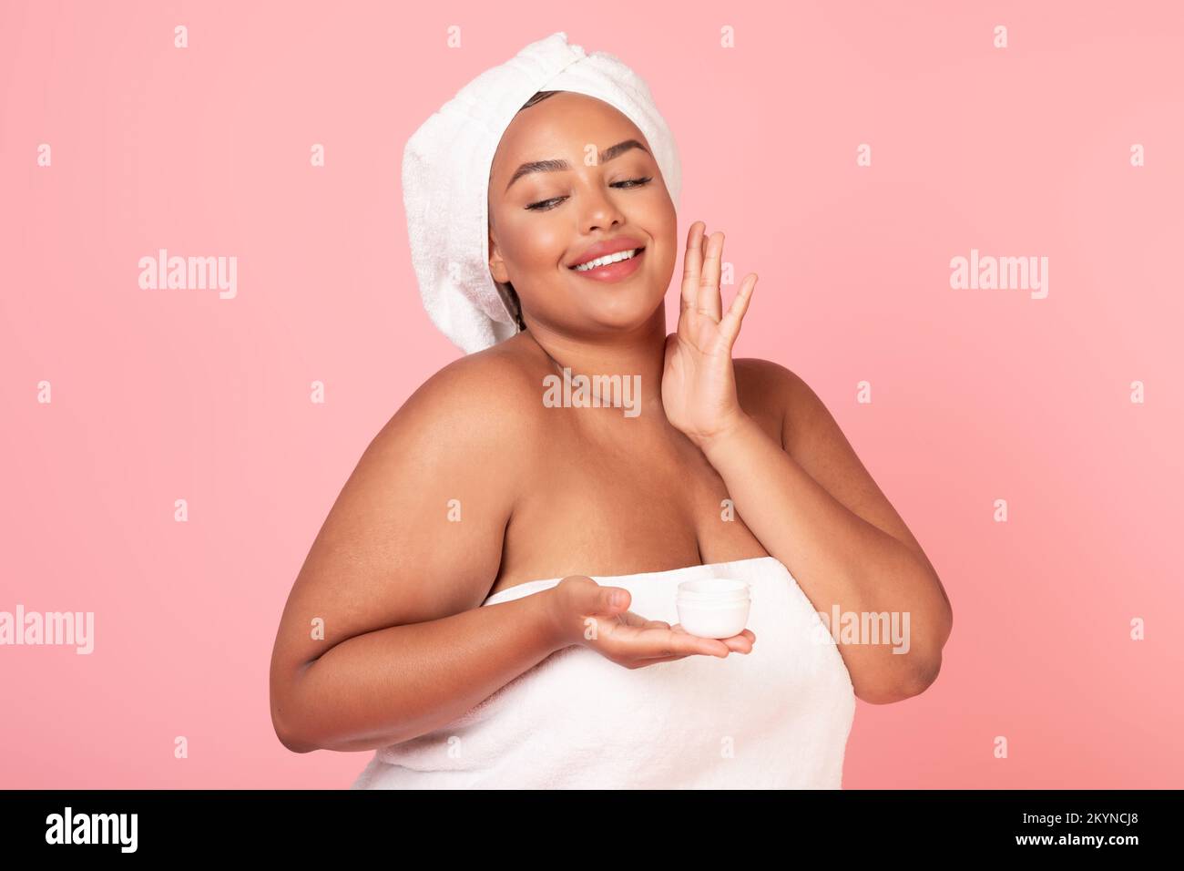 Skin care concept. Happy black oversize lady with towel on head applying moisturizing face cream, pink background Stock Photo