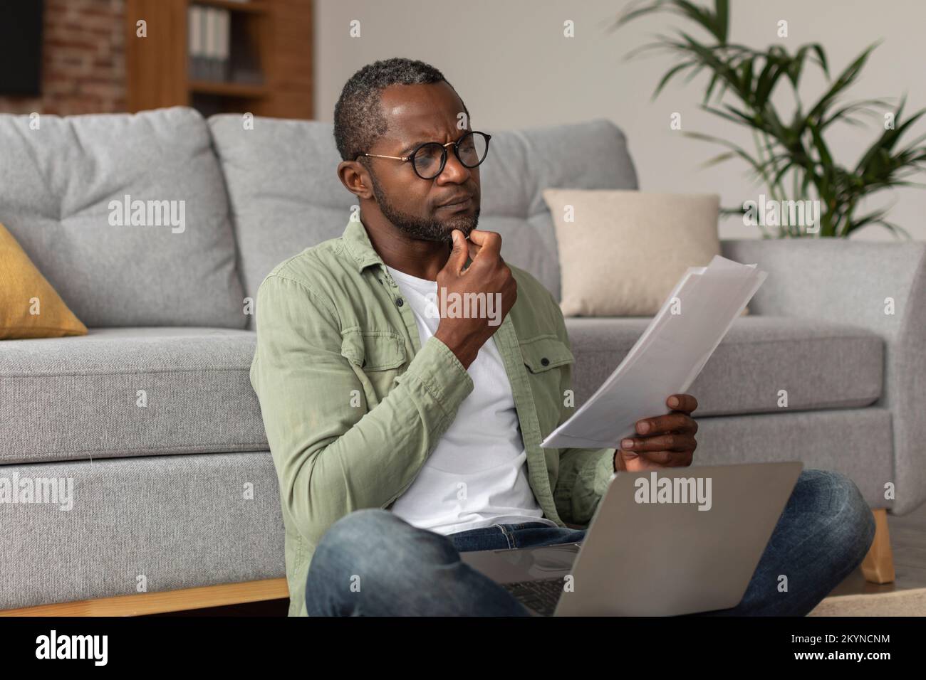 Concentrated pensive middle aged african american guy in glasses with laptop reads document Stock Photo