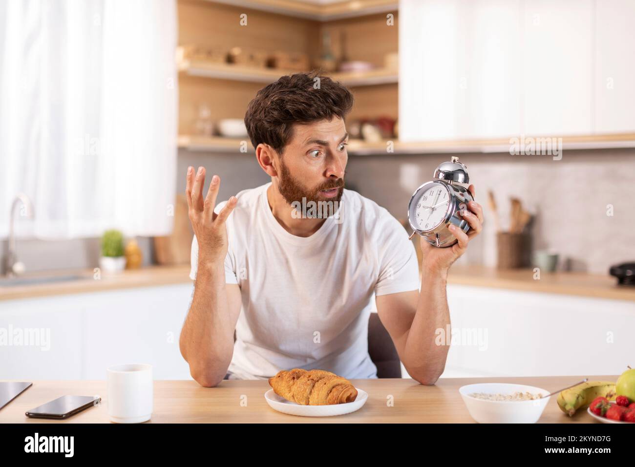 Shocked sad adult caucasian man with beard in white t-shirt looks at alarm clock and late to meet or work Stock Photo