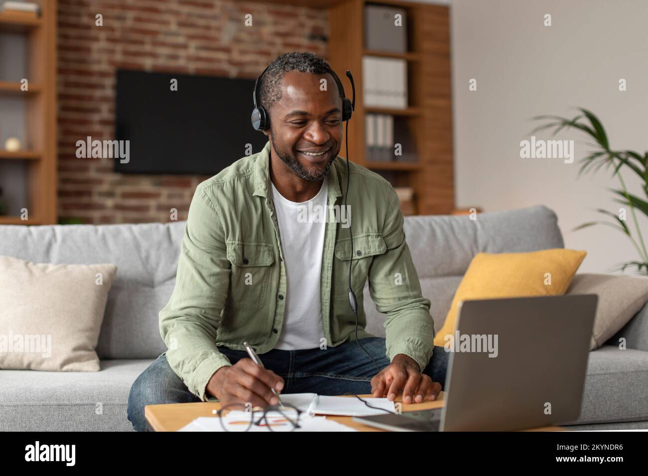 Smiling middle aged african american guy in headphones looks at laptop, makes notes in living room interior Stock Photo