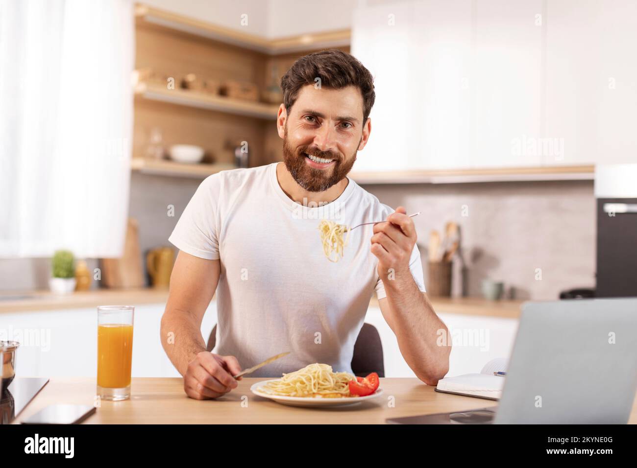 Happy handsome mature caucasian bearded male eating pasta in minimalist kitchen interior with laptop Stock Photo
