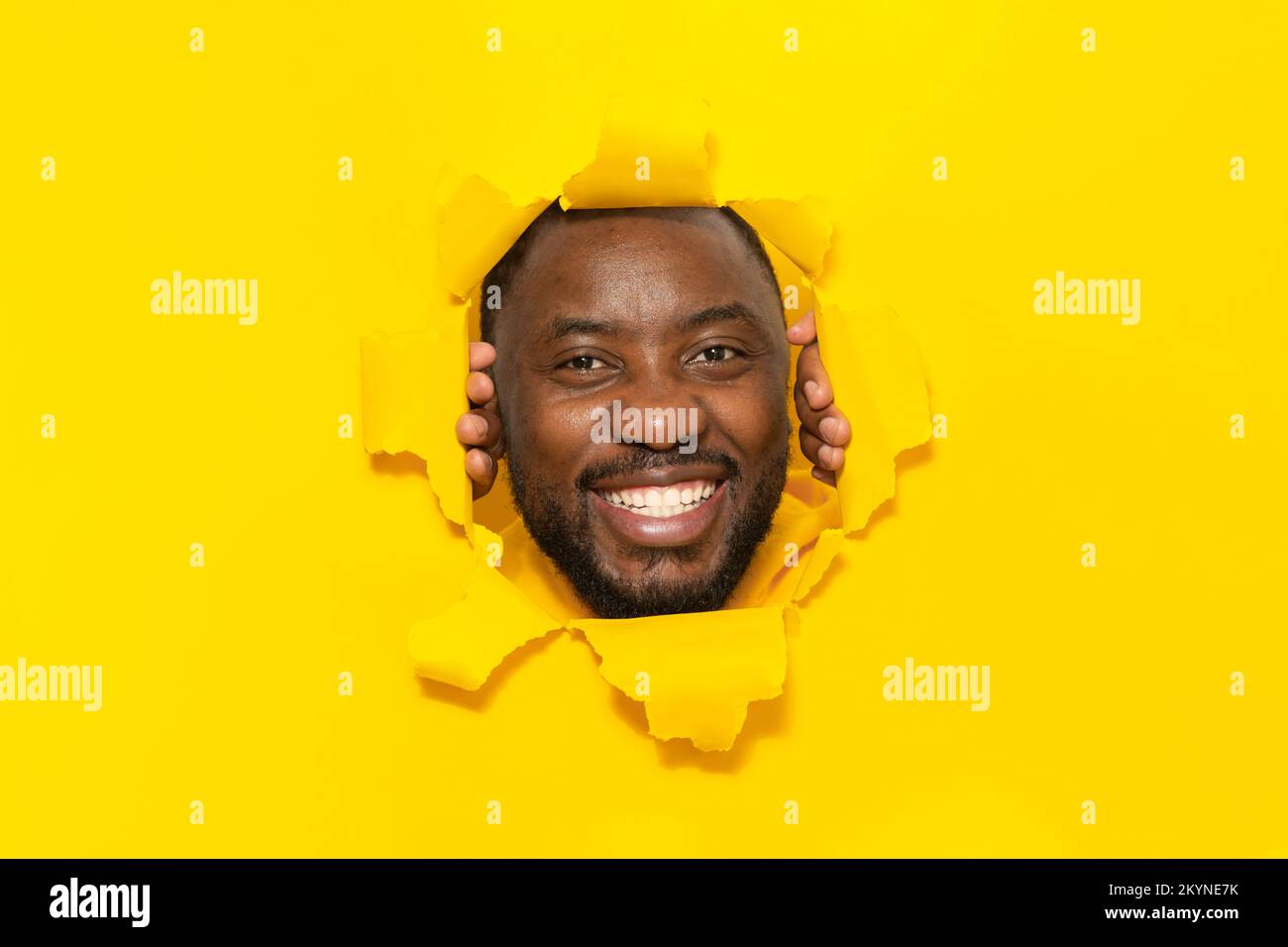 Happy african american man posing in torn yellow paper, smiling looking at camera through hole on studio background Stock Photo