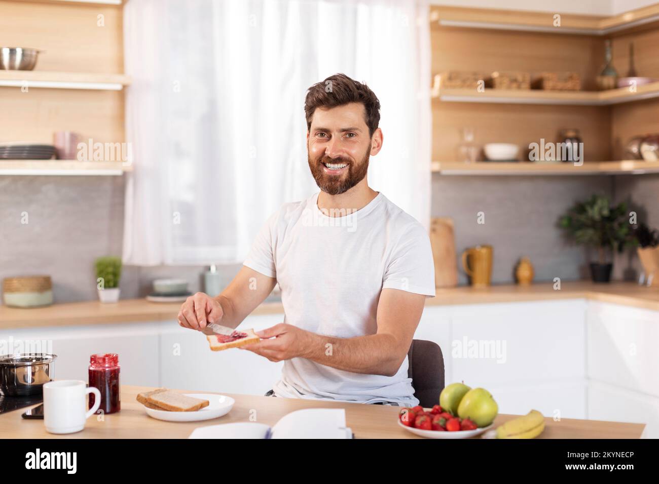 Cheerful middle aged caucasian male with beard in white t-shirt makes sandwich with fruits for breakfast Stock Photo