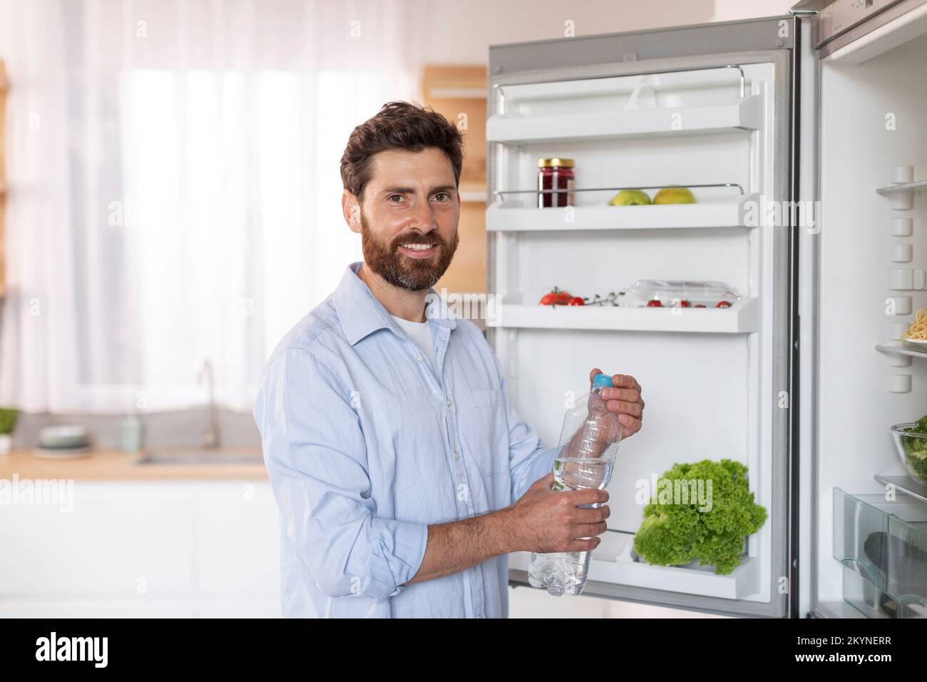 Cheerful mature caucasian male with beard in shirt opens refrigerator door, takes bottle of water in kitchen interior Stock Photo
