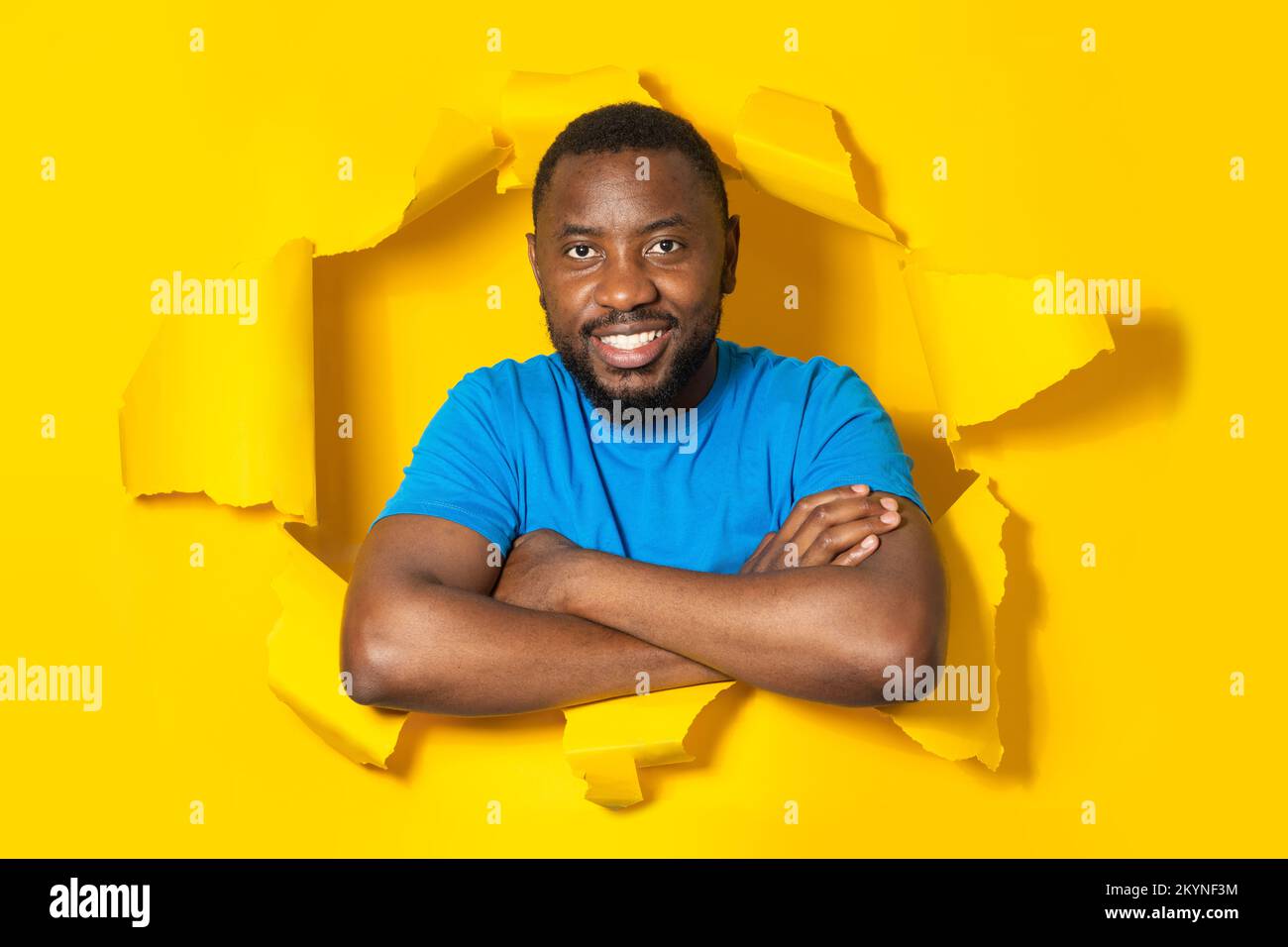 Portrait of positive black man posing with folded arms in torn paper hole, looking and smiling at camera Stock Photo