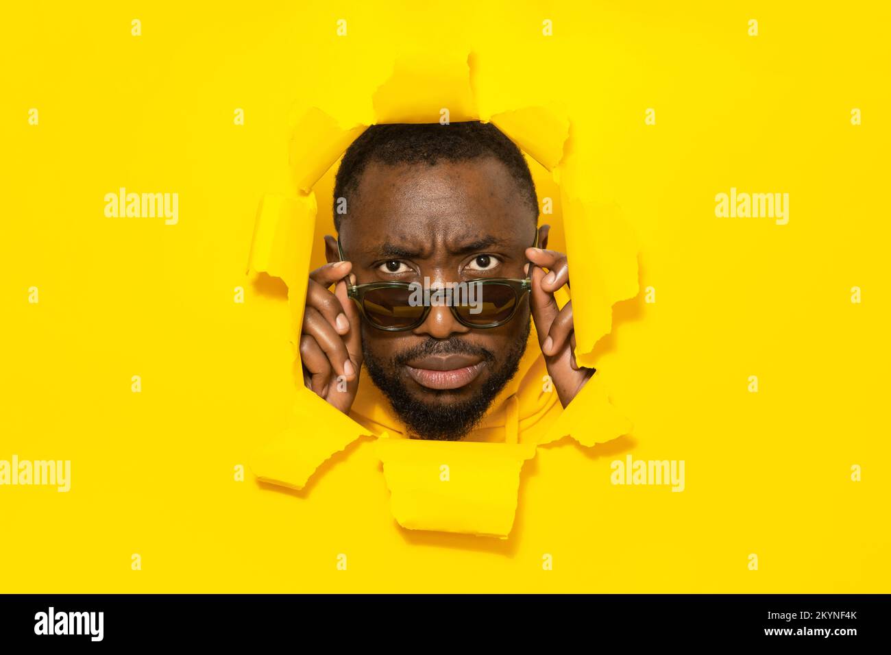 Serious african american man looking at camera through sunglasses, breaking through yellow paper background Stock Photo