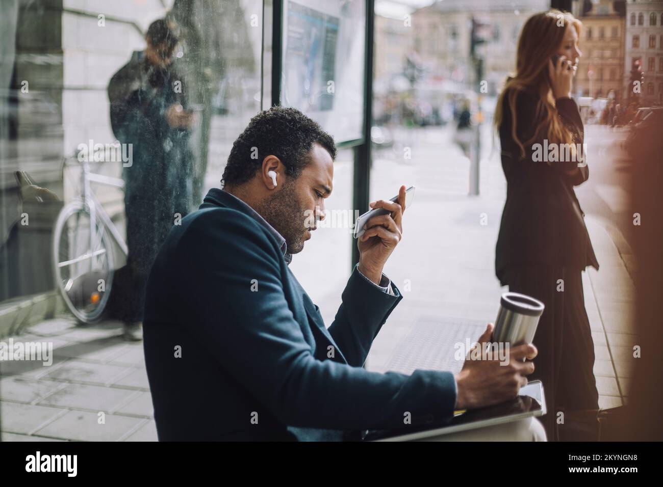 Side view of male professional sending voicemail while sitting against businesswoman talking on mobile phone at bus stop Stock Photo